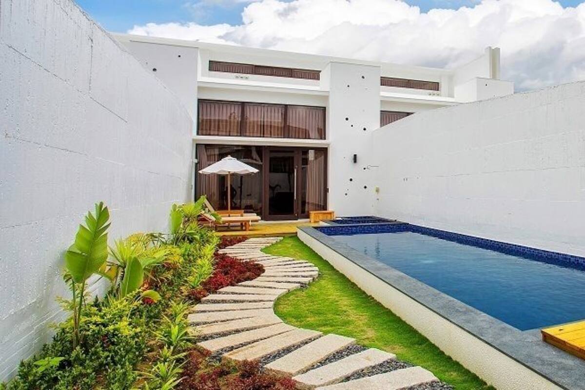 Un área de piscina moderna al aire libre con piscina azul cristalina, terraza de madera y zona de estar con mesa y sillas.