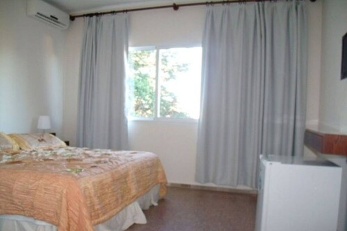 A bedroom with a bed, a window with curtains, a bedside table with a lamp, and a white wardrobe.