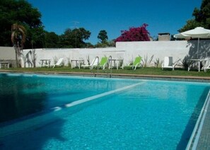 Una piscina con agua cristalina azul, rodeada de tumbonas y un muro blanco con un arbusto rosado.