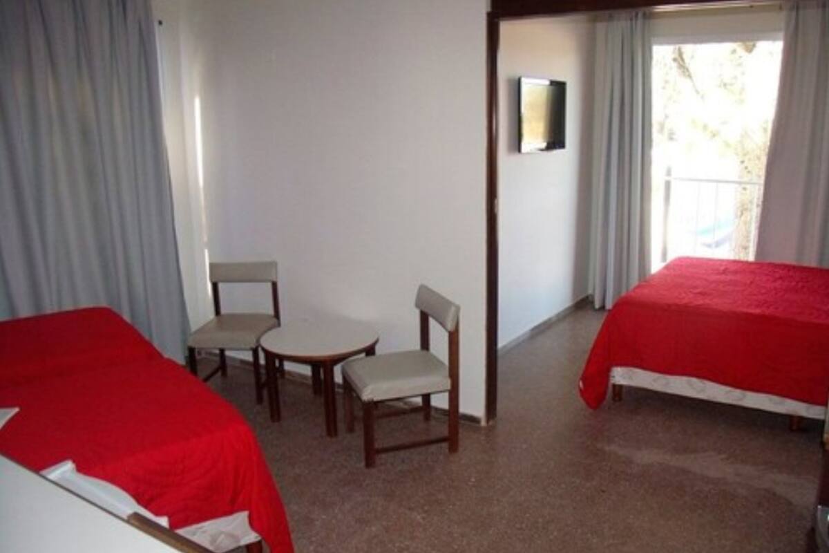 A hotel room with two single beds, a small table with two chairs, a mirror on the wall, and a window with curtains.