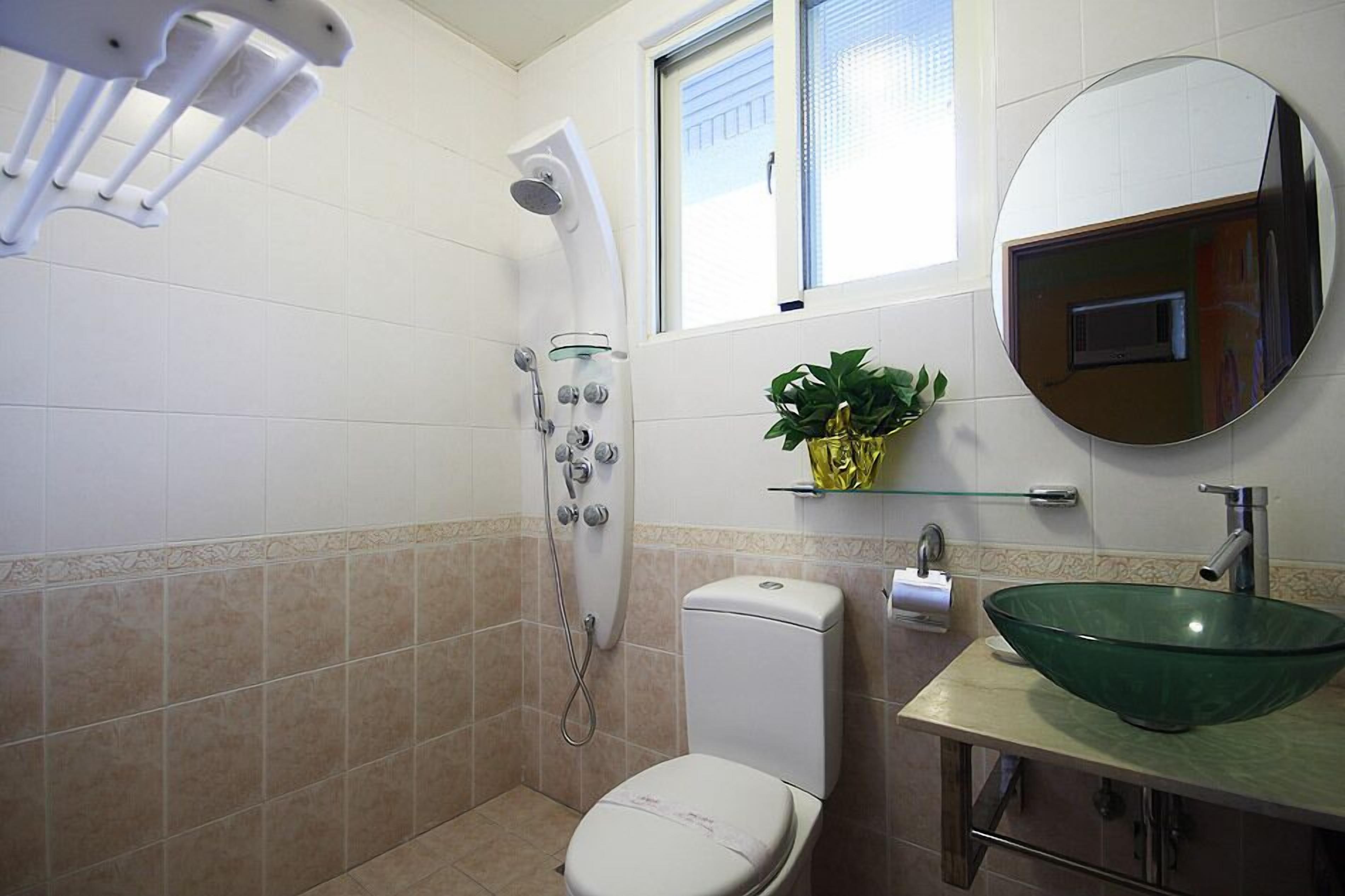 Une salle de bain avec un lavabo en verre, des toilettes, une douche avec un pommeau de douche amovible et un miroir intégrant un écran de télévision.