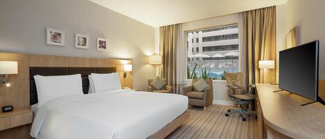 A hotel room with a large bed, a desk with a chair, a television, and a window with a view of a building and umbrellas.