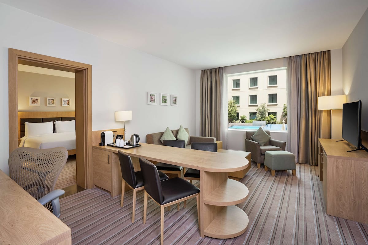A hotel room with a bed, a desk, a dining table, a sofa, a TV, and a window with a view of buildings.