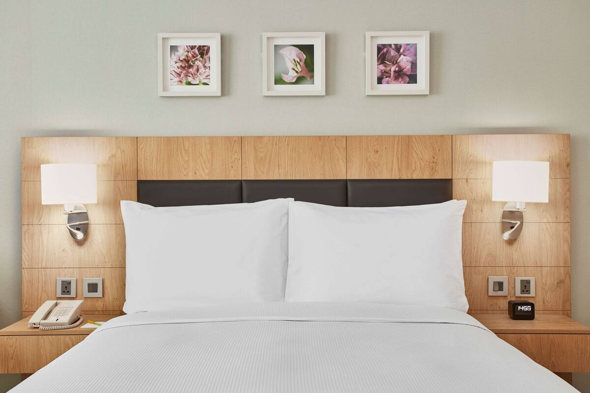 A hotel room with a wooden headboard, white bedding, bedside lamps, a clock, and three framed floral pictures on the wall.