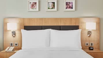 Habitación de hotel con cabecera de madera, ropa de cama blanca, lámparas de noche, un reloj y tres cuadros con motivos florales en la pared.