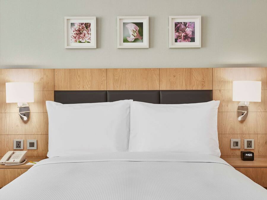 Habitación de hotel con cabecera de madera, ropa de cama blanca, lámparas de noche, un reloj y tres cuadros con motivos florales en la pared.