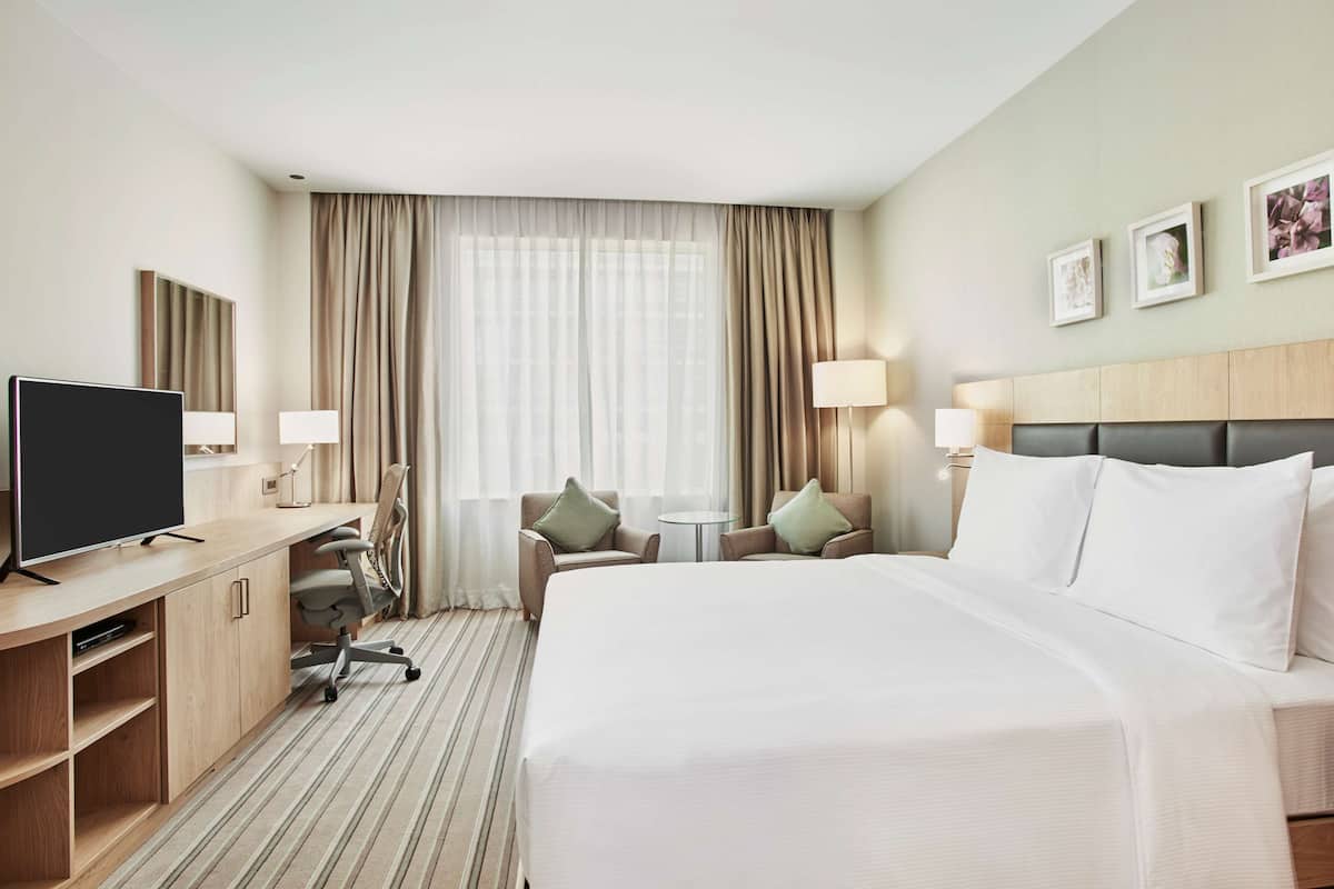 A hotel room with a large bed, a desk with a TV, two armchairs, a small table, and a window with curtains.