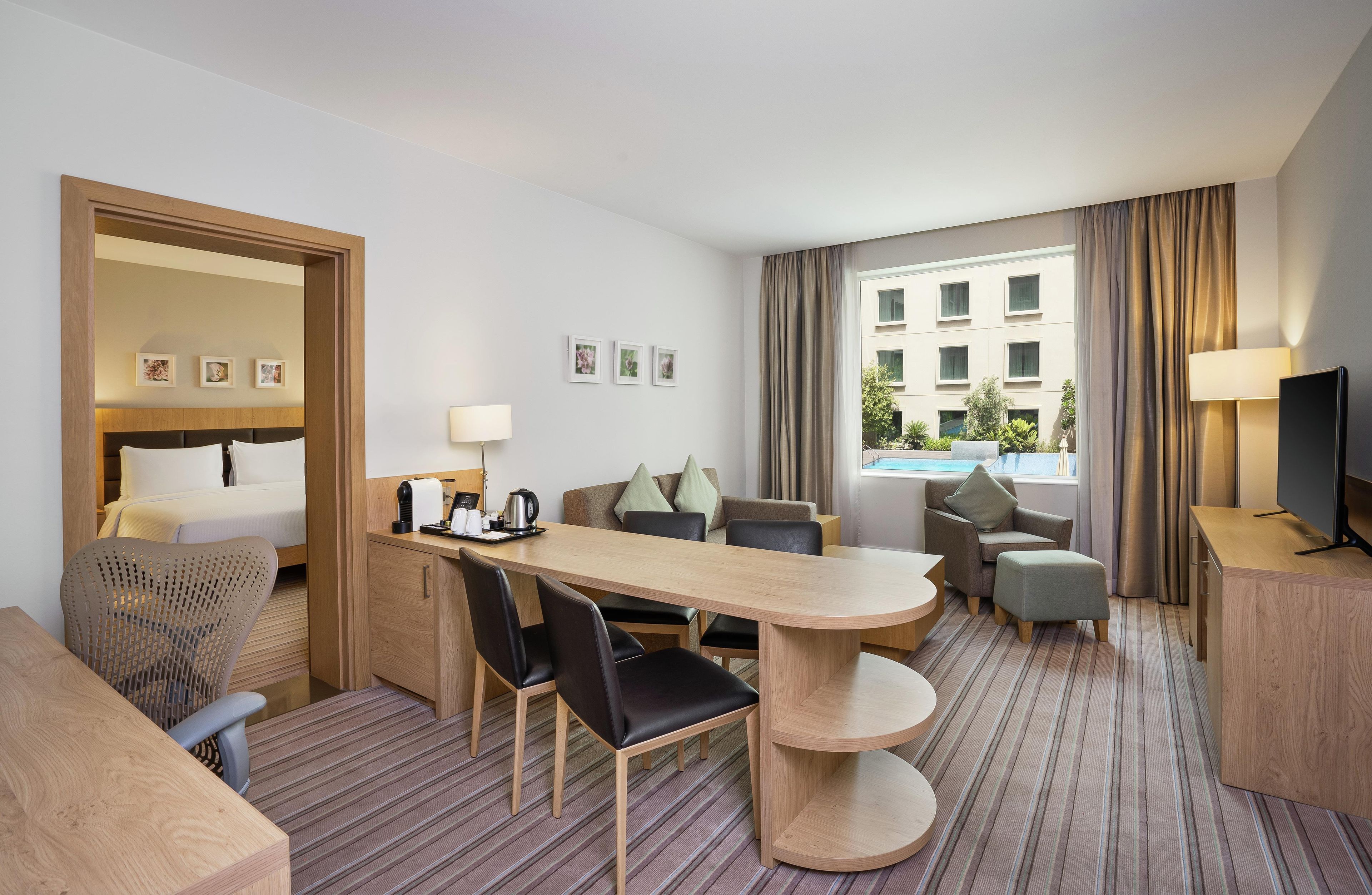 Ein Hotelzimmer mit Bett, Schreibtisch, Esstisch, einer Couch, einem Fernseher und einem Fenster mit Vorhängen.