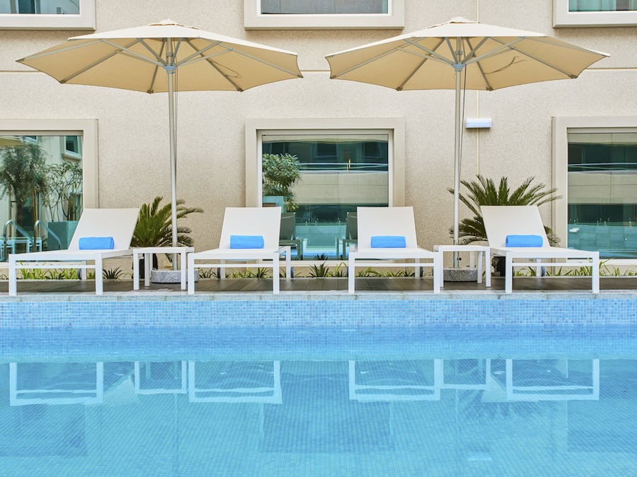 Zona junto a la piscina con sillones de descanso y sombrillas, una piscina cristalina y un edificio con grandes ventanales al fondo.