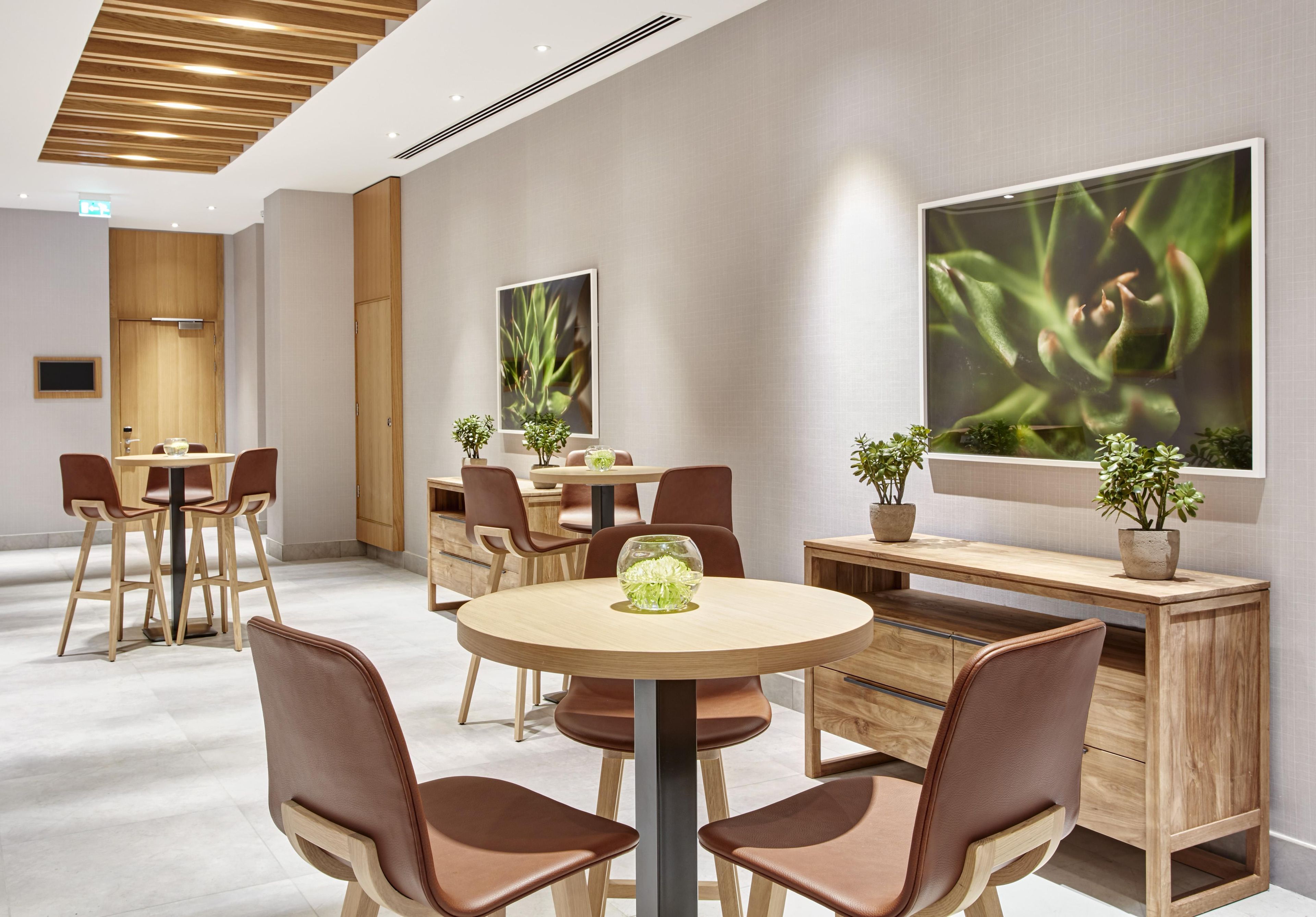 A modern hotel room with a round dining table, two brown leather chairs, a wooden desk with a plant, and a large window with a plant view.
