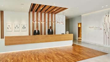 A reception area with a wooden reception desk, two staff members, and a wall with Arabic text.