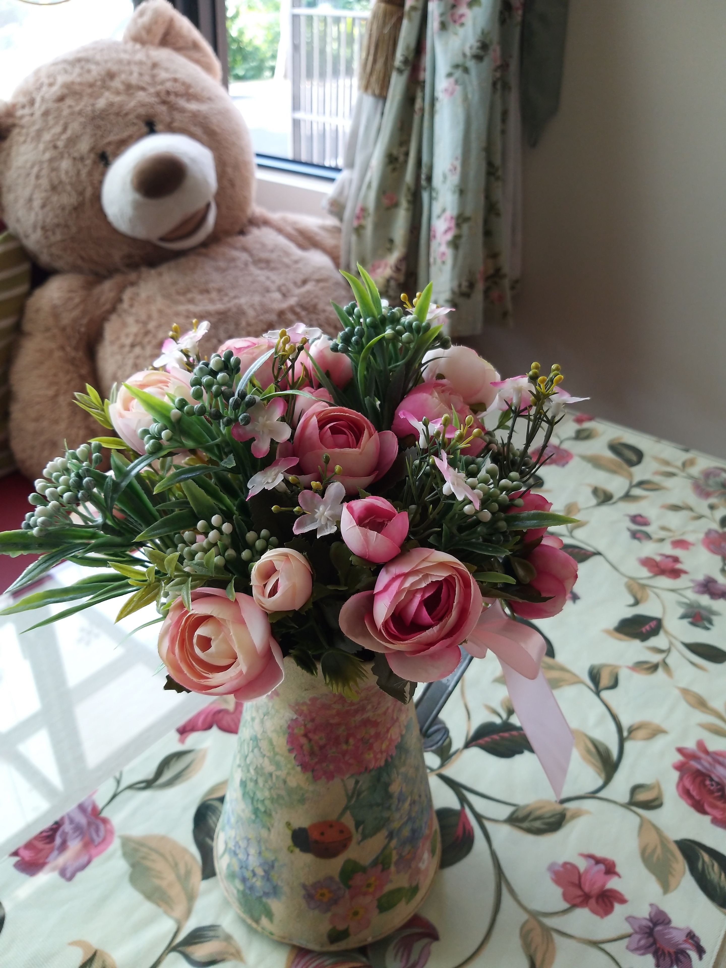 A floral arrangement in a vase on a table with a floral tablecloth, a teddy bear in the background, and a window with curtains.