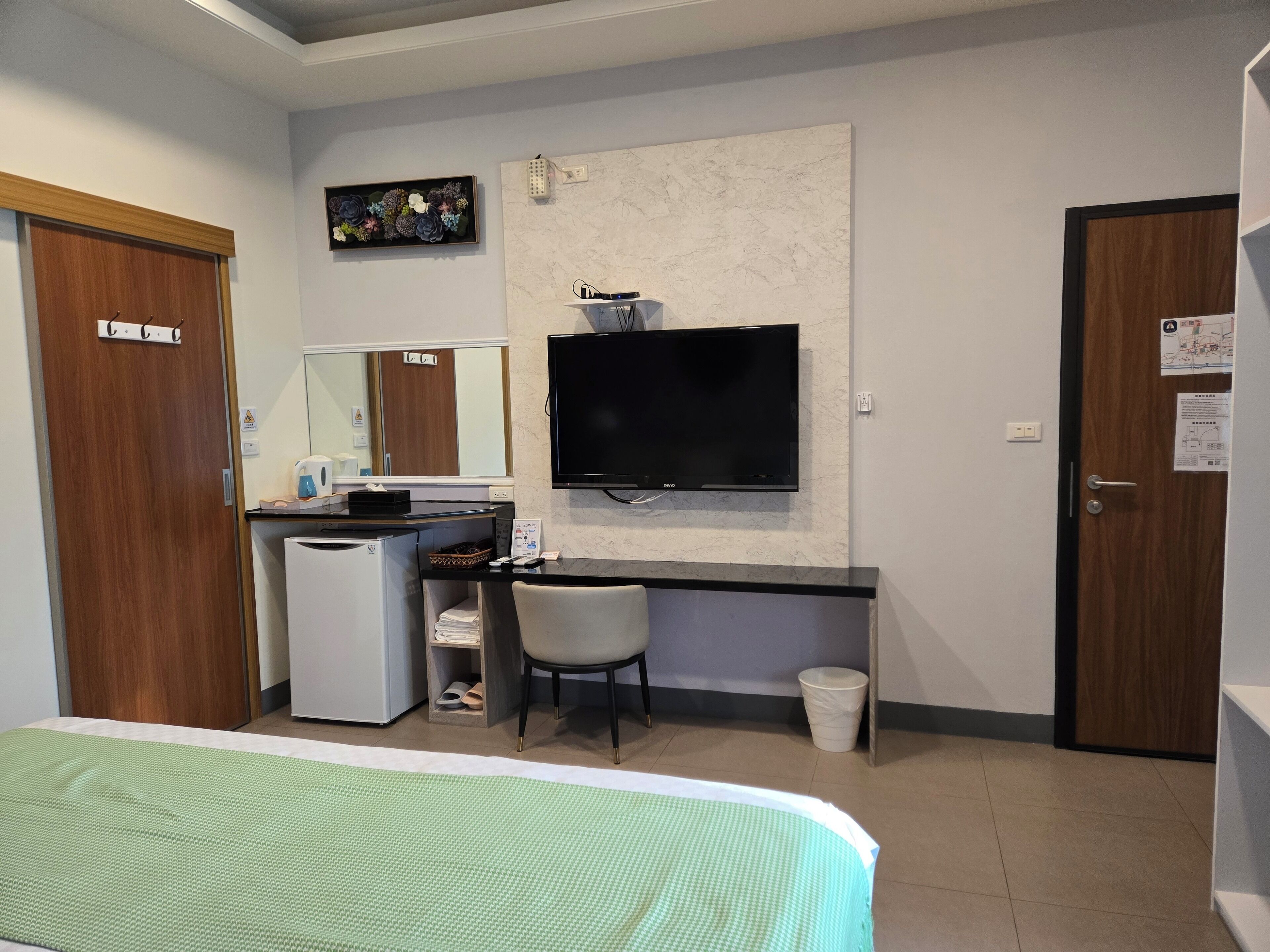 A hotel room with a bed, a desk with a computer, a television, a small refrigerator, a trash bin, and a mirror.