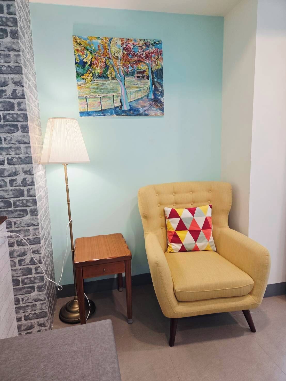 Una habitación con un sillón amarillo, una pequeña mesita de madera, una lámpara de pie y un cuadro llamativo en la pared.
