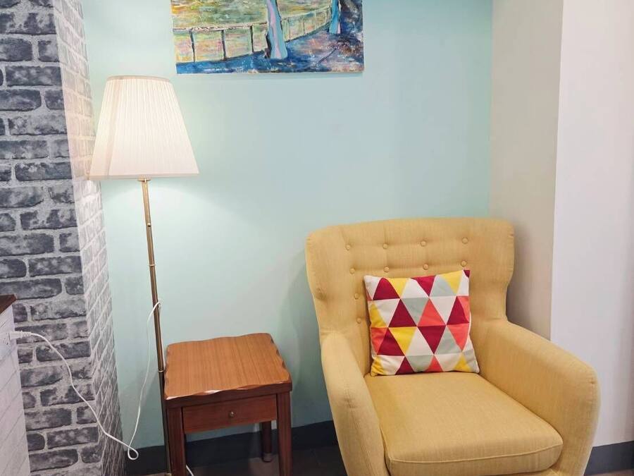 Una habitación con un sillón amarillo, una pequeña mesita de madera, una lámpara de pie y un cuadro llamativo en la pared.