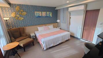 A hotel room with a bed, a sofa, a small table, and a desk. There is a painting on the wall and an air conditioning unit above the bed.