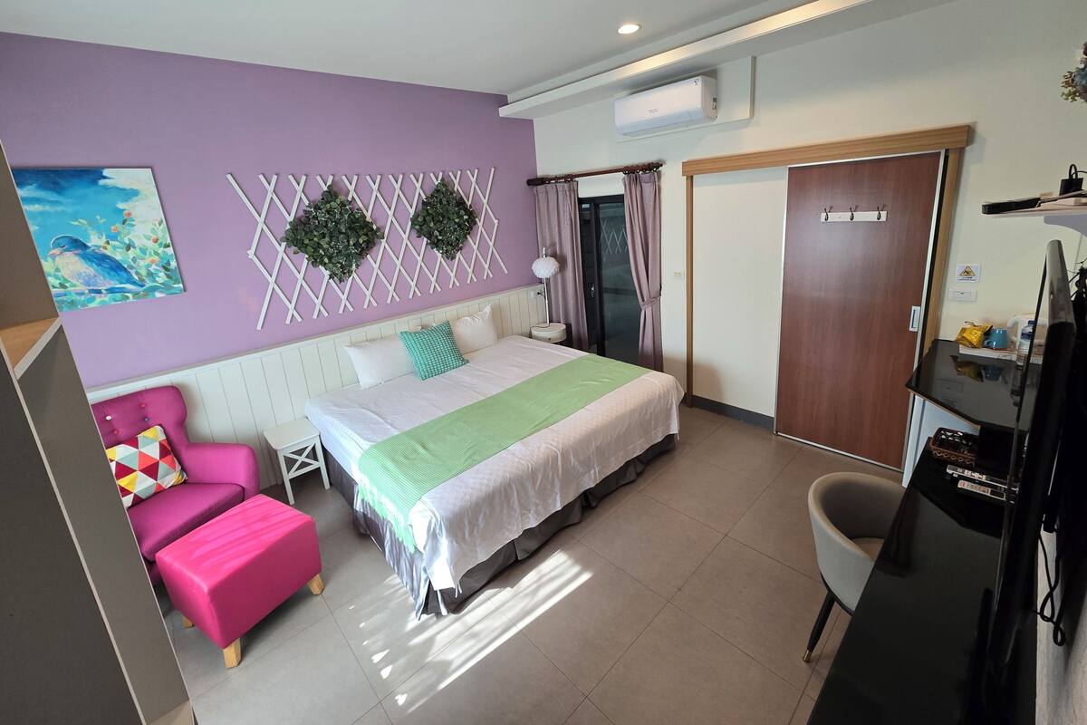A bedroom with a bed, a pink chair, a desk, and a TV.