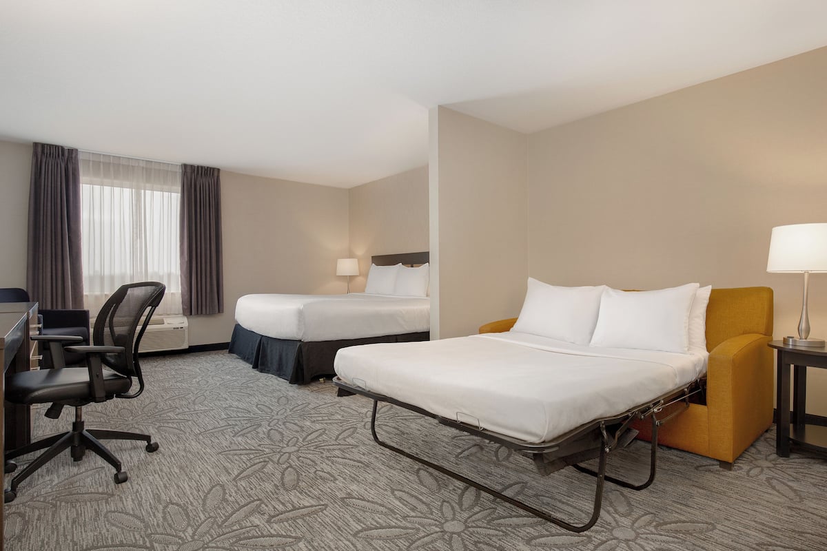 A hotel room with a single bed, a desk with a chair, a second bed, a small table with a lamp, and a window with curtains.