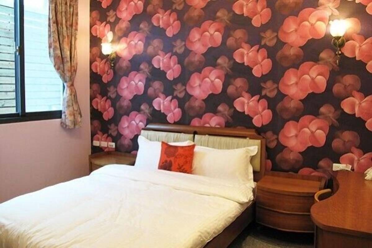 A bedroom with a floral wallpaper, a bed with white bedding, a nightstand, and a wooden dresser.