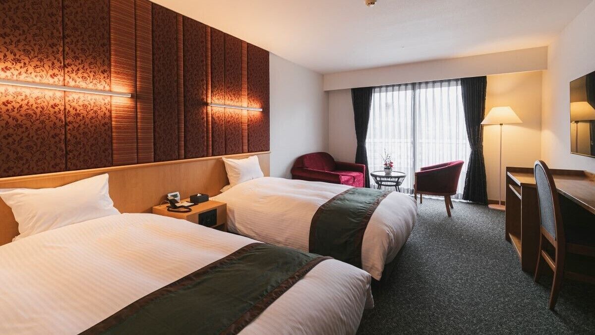 Een hotelkamer met twee bedden, een bureau, een stoel, een kleine tafel en een groot raam met gordijnen.