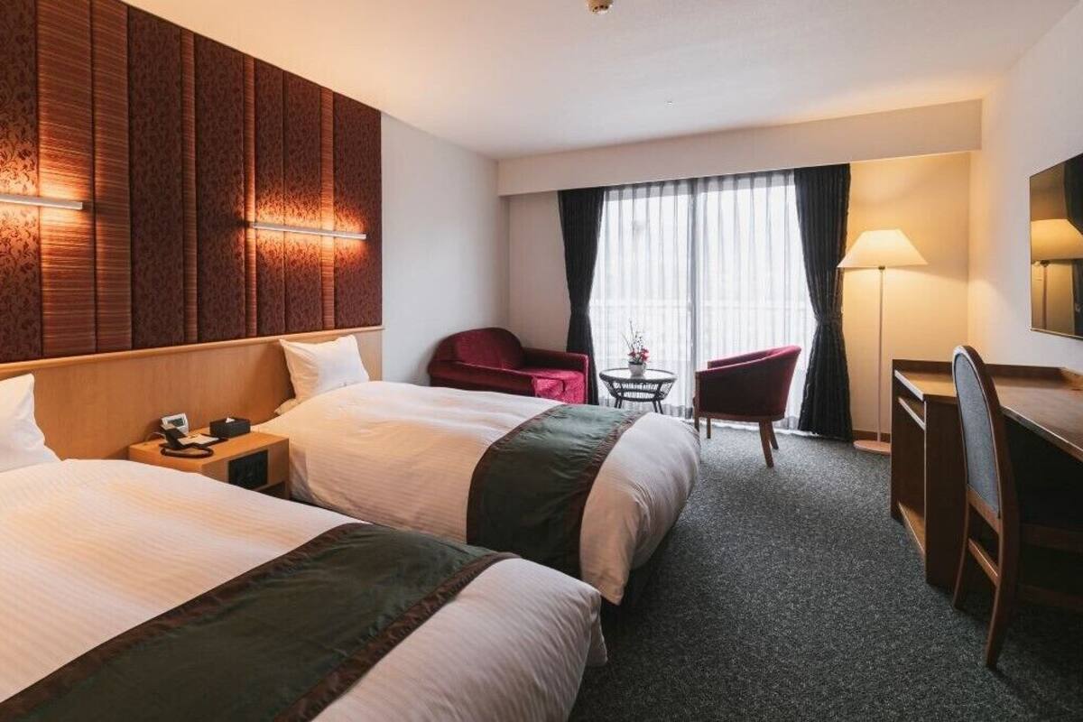 A hotel room with two beds, a desk, a chair, a small table, and a large window with curtains.