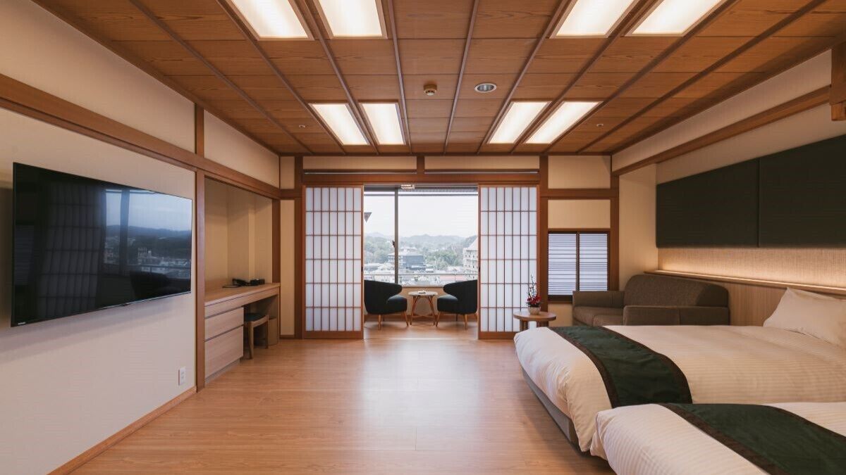 Een hotelkamer met een groot bed, een televisie, een bureau met stoelen, en uitzicht op de stad door grote ramen.