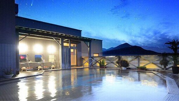 Zona de piscina con terraza de madera, plantas en macetas y un edificio con ventanas iluminadas.