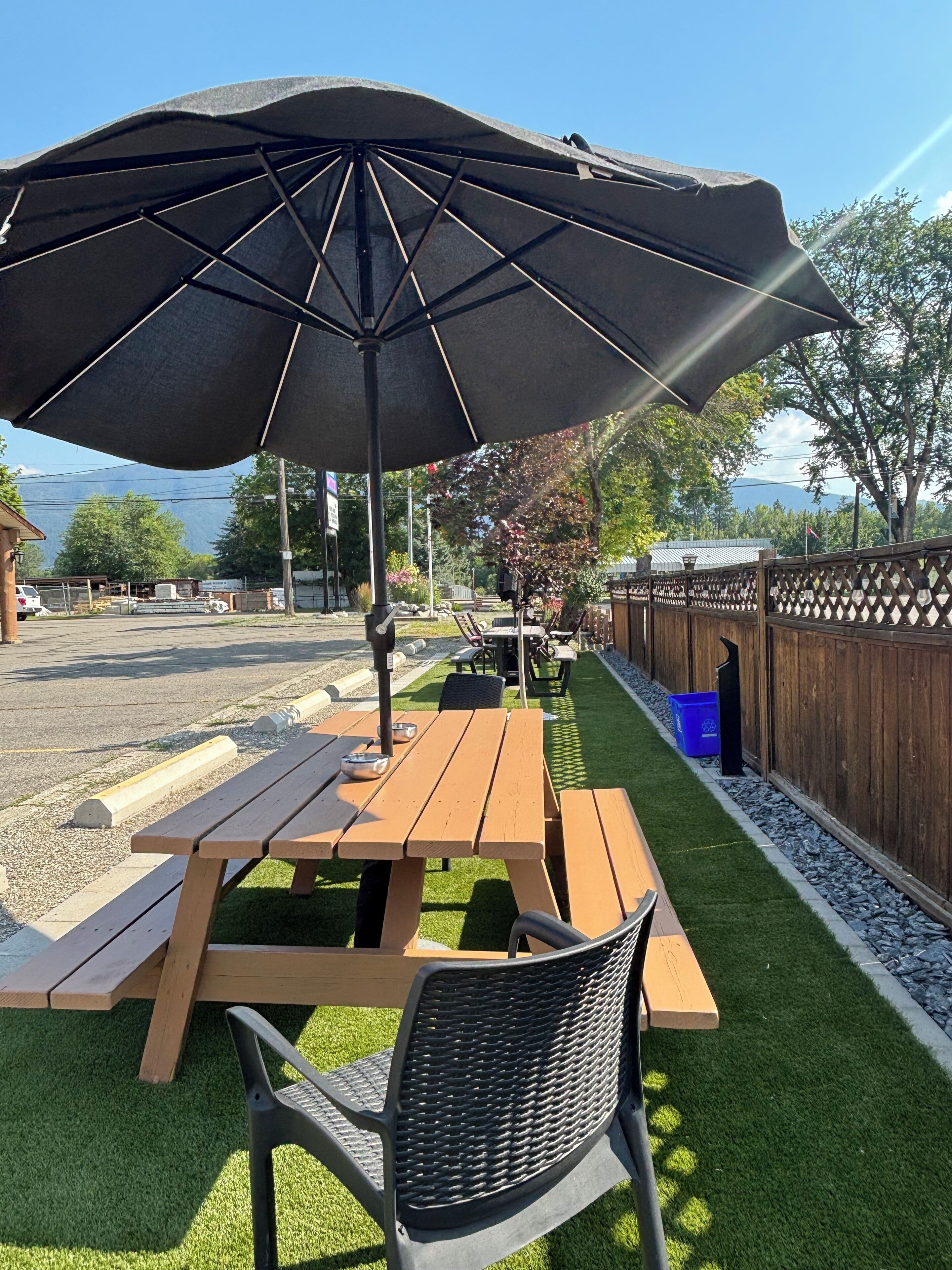 An outdoor seating area with wooden picnic tables and benches, a black umbrella, and a fenced garden.