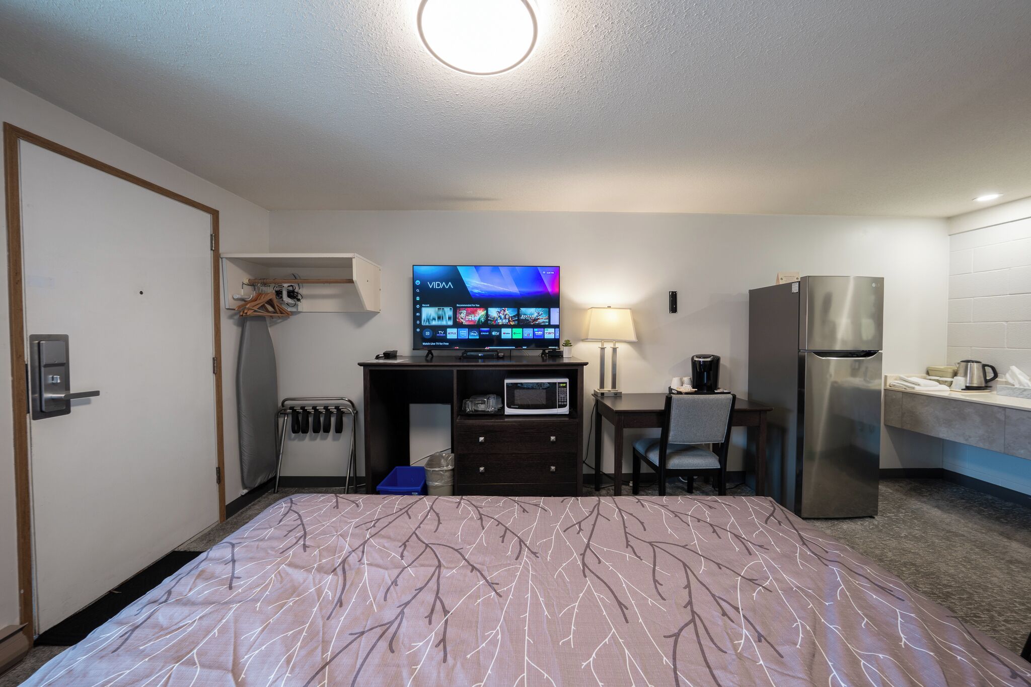 A hotel room with a bed, a desk with a TV, a microwave, a refrigerator, and a small kitchenette.