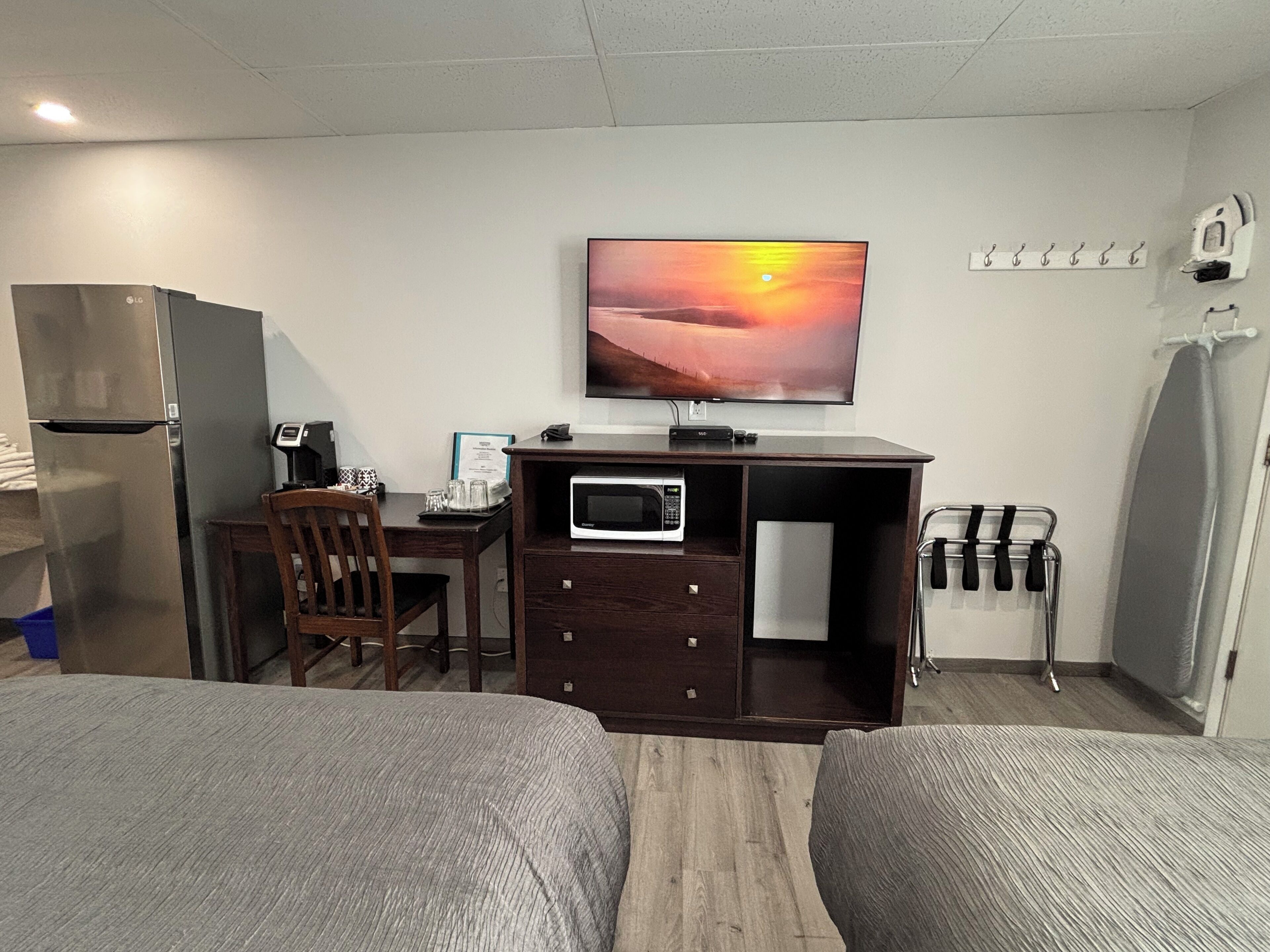 A hotel room with a bed, a desk with a computer, a microwave, a refrigerator, a TV, and a window with a sunset view.