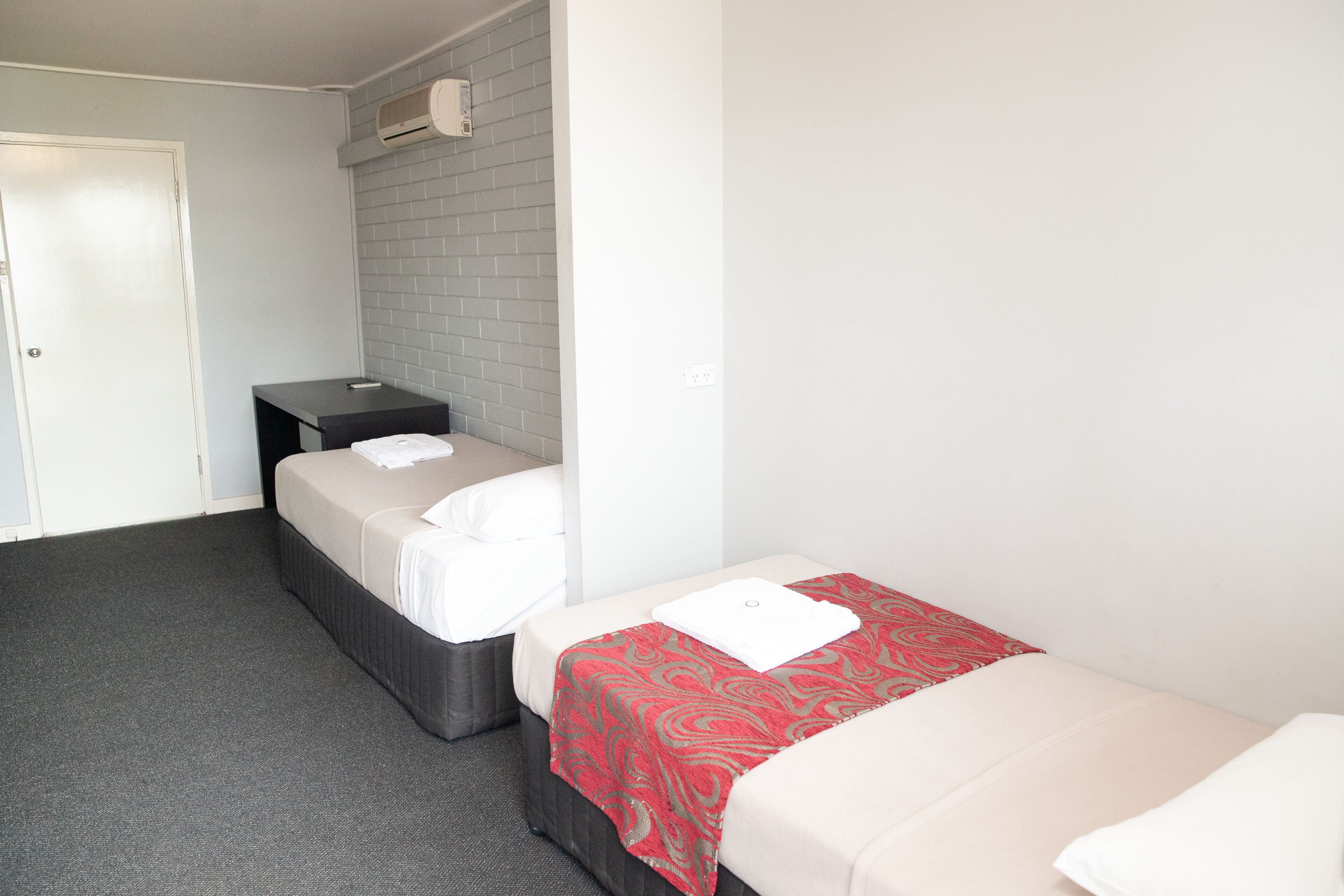 Een kamer met twee bedden, een klein bureau, een witte deur en een grijze bakstenen muur.
