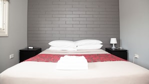 A neatly made bed with white linens and a red patterned throw, two bedside tables with lamps, and a grey brick wall. - Royal Hotel Singleton (Singleton)