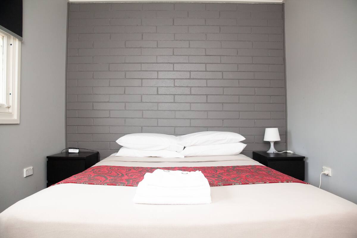 A neatly made bed with white linens and a red patterned throw, two bedside tables with lamps, and a grey brick wall.