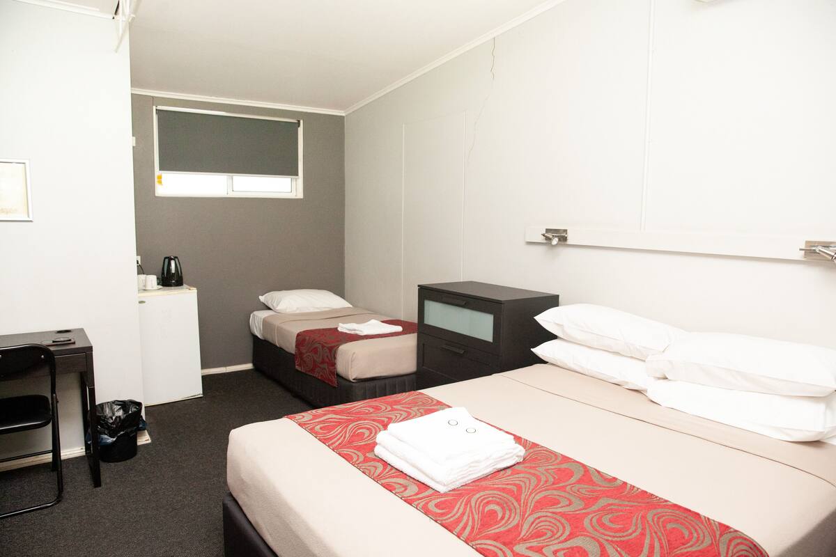 A hotel room with a bed, a desk, a chair, a small table, a nightstand, and a window.
