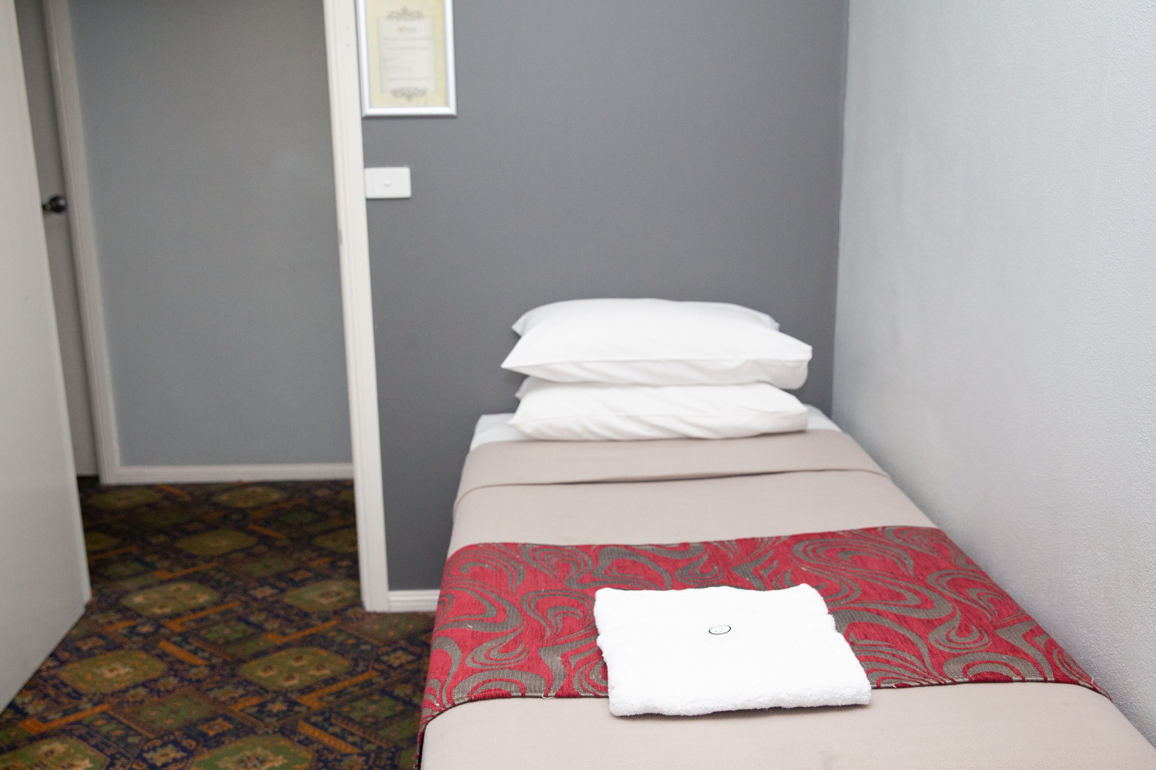 A small, narrow hotel room with a single bed, a patterned rug, and a framed certificate on the wall.