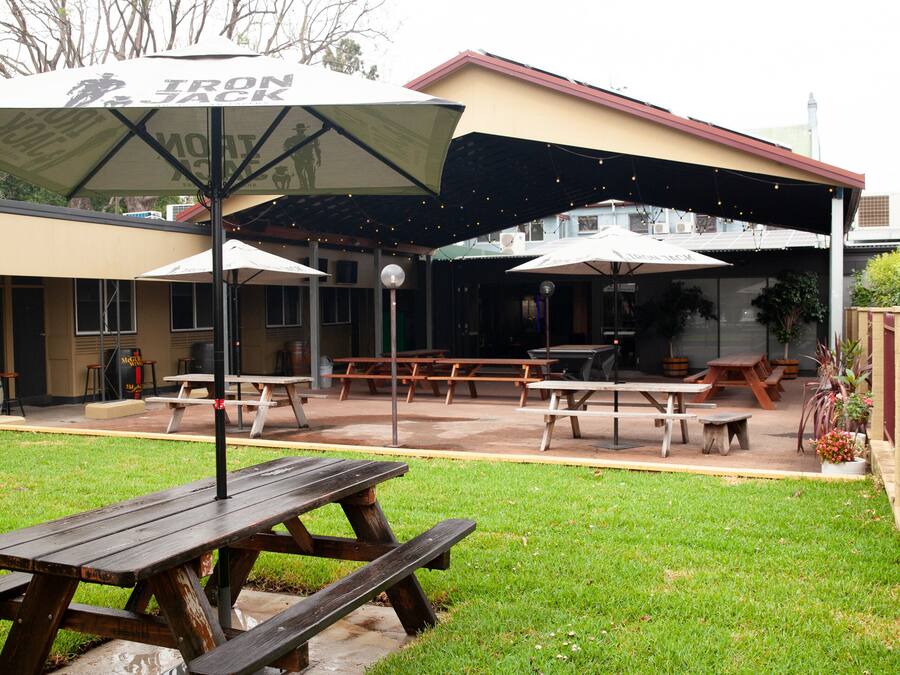 An outdoor patio area with picnic tables and umbrellas, surrounded by a grassy lawn and a building with a sloped roof.
