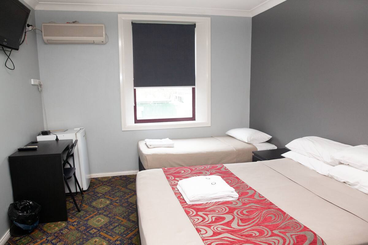 A hotel room with a bed, a desk, a chair, a small table, an air conditioner, and a window with a blind.