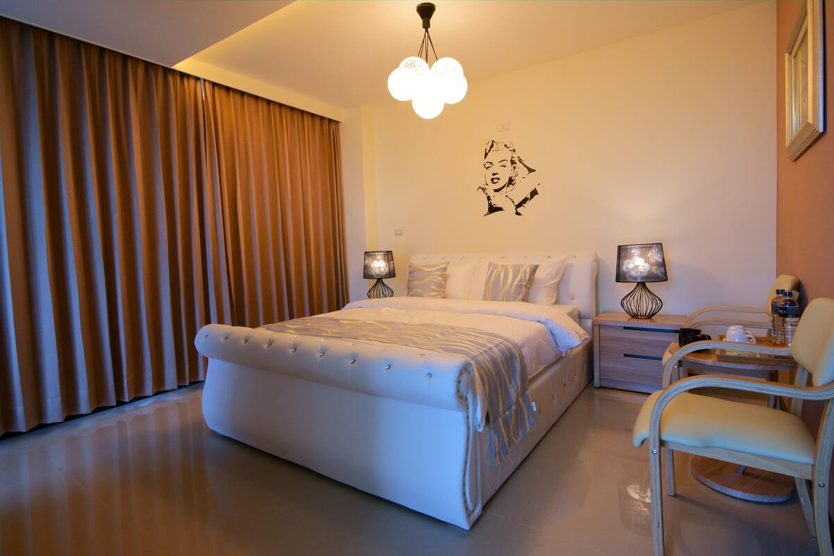 A modern bedroom with a large bed, bedside lamps, a desk with a chair, and a wall decoration.