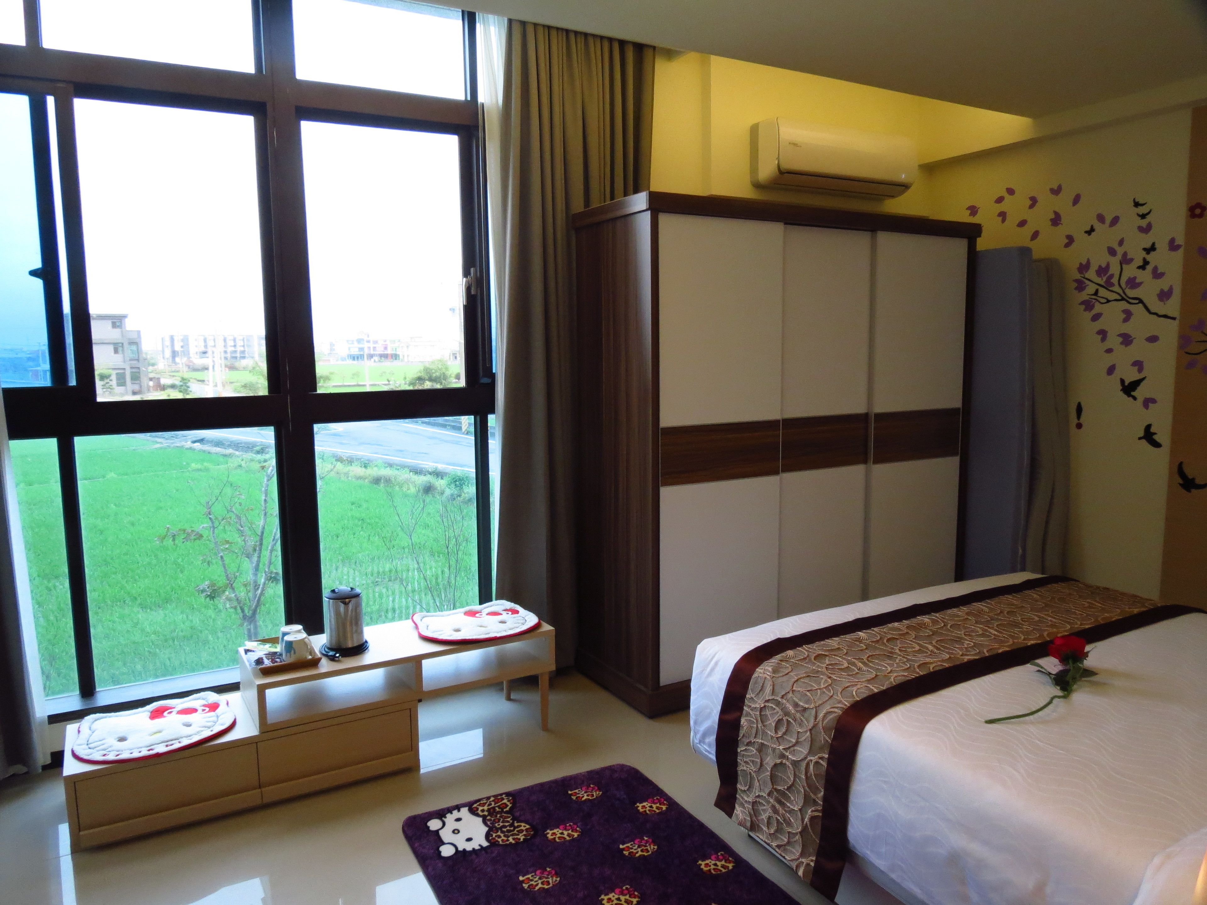 Habitación de hotel con un ventanal grande, un armario, una mesita de noche con un tapete de Hello Kitty, una cama con una rosa y vistas a un campo verde y edificios.
