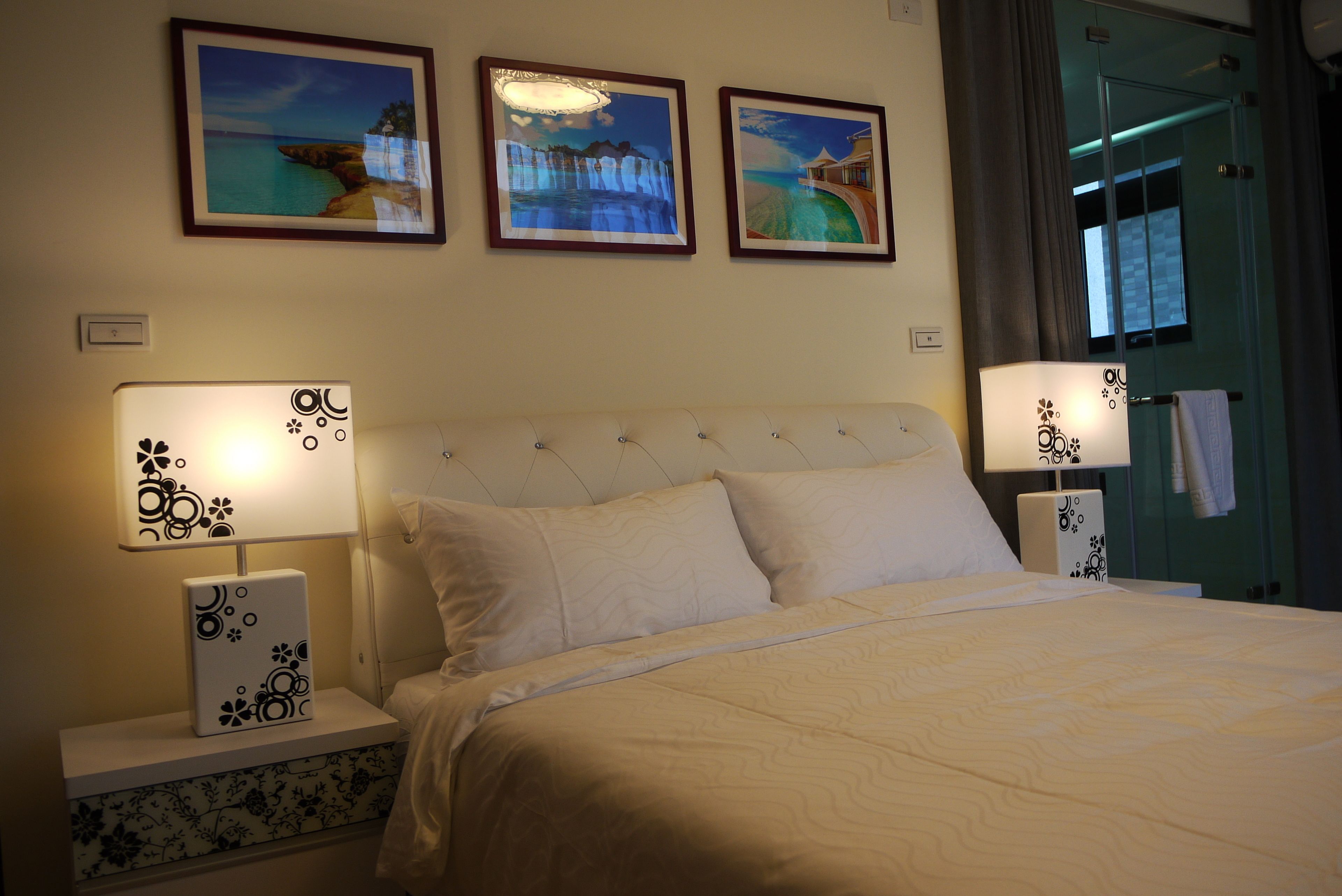 Una cama bien hecha con ropa de cama blanca, dos lámparas de noche con diseños florales y tres cuadros en la pared que representan escenas costeras.