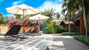 A sunlit outdoor seating area with lounge chairs and umbrellas, surrounded by lush tropical plants and a brick building. - Royal Lee Resort & Spa (Sa Khu)