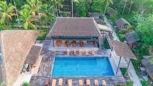Vista aérea de un resort con una piscina central rodeada de sillas de madera para tomar el sol, un edificio con techo de paja y exuberante vegetación tropical.