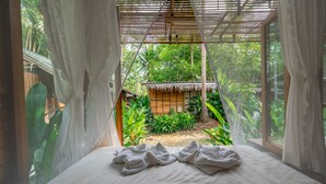 Habitación con cama, cortinas blancas y vista a una construcción de techo de paja rodeada de vegetación.
