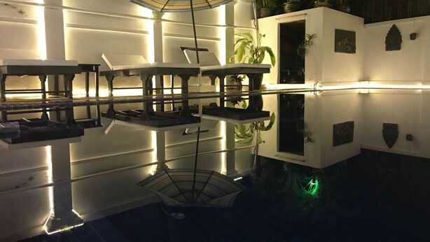 Ein spiegelnder Pool mit Tischen und Stühlen, umgeben von Mauern mit kleinen Fenstern und Topfpflanzen.