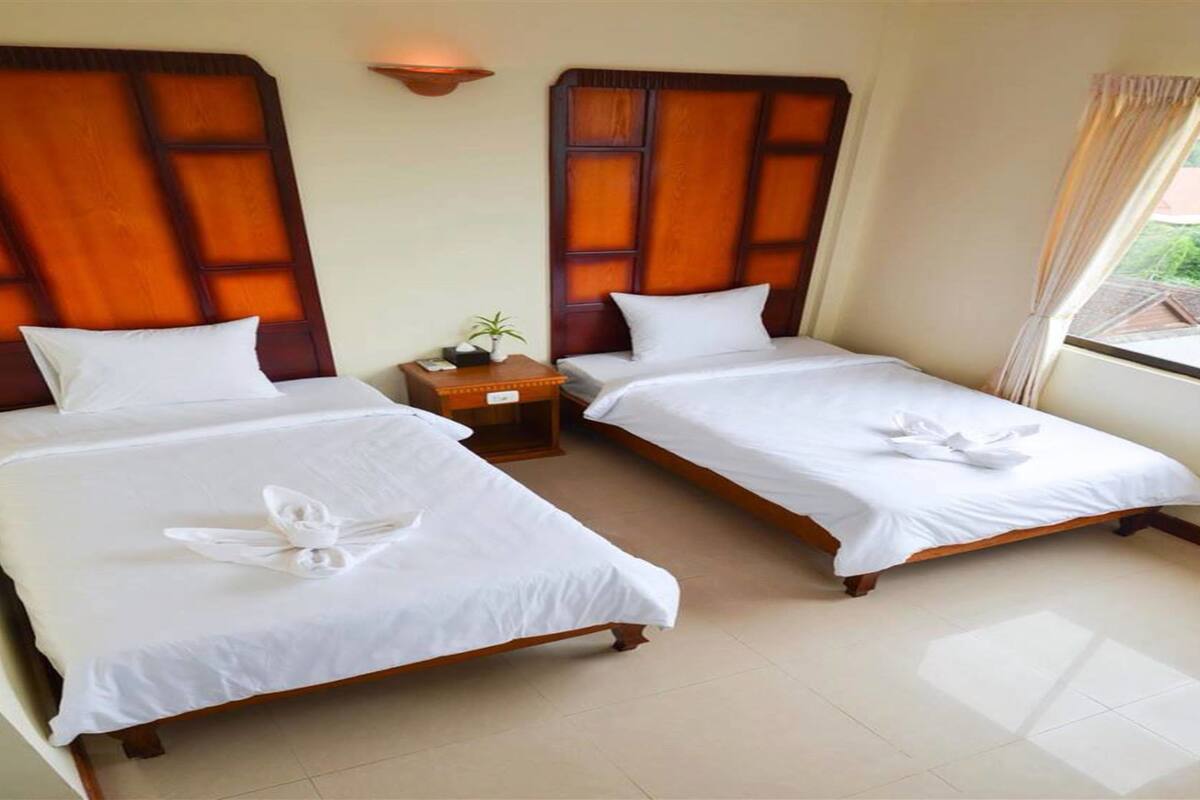 Ein Hotelzimmer mit zwei Einzelbetten, Holz-Kopfteilen, weißen Laken, einem kleinen Nachttisch mit Pflanze und einem Fenster mit Vorhängen.