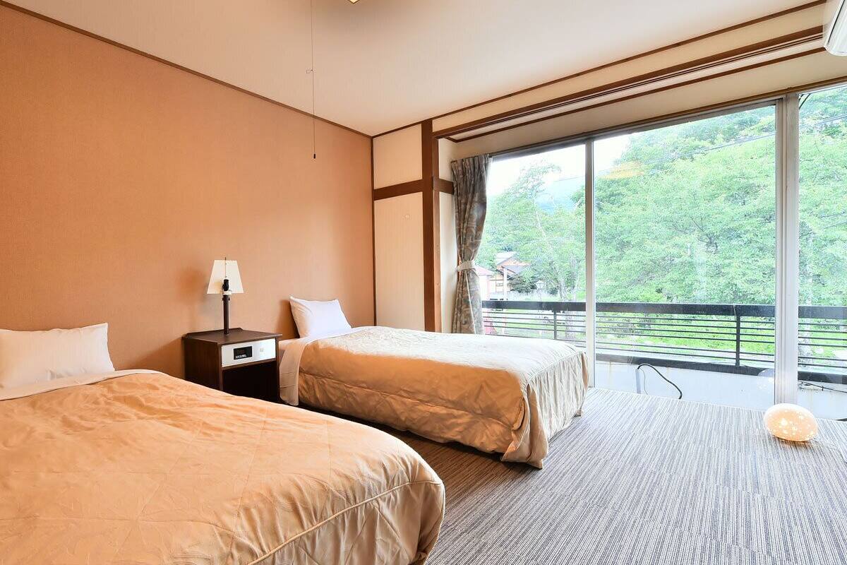 A hotel room with two beds, a nightstand, a lamp, and a large window with a view of greenery.