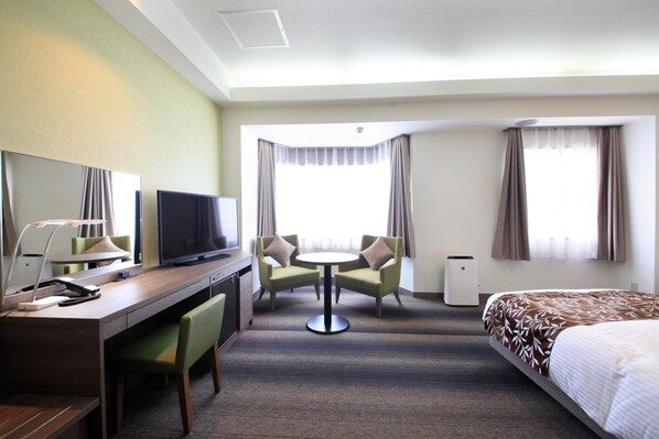A hotel room with a bed, a desk with a TV, a chair, a small table, and a window with curtains.