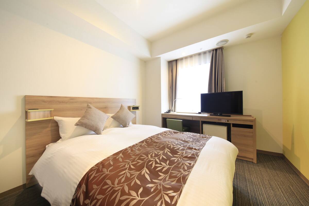 A hotel room with a bed, a desk with a television, and a window with curtains.
