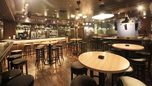 Zona de bar con taburetes, mesas y mostrador que exhibe botellas y vasos.