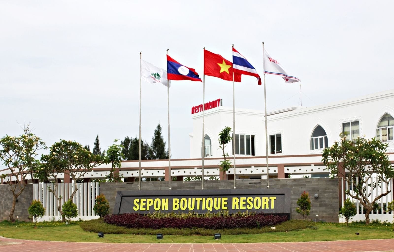 Um edifício com bandeiras, uma placa com a inscrição "SEPON BOUTIQUE RESORT" e um jardim bem cuidado.